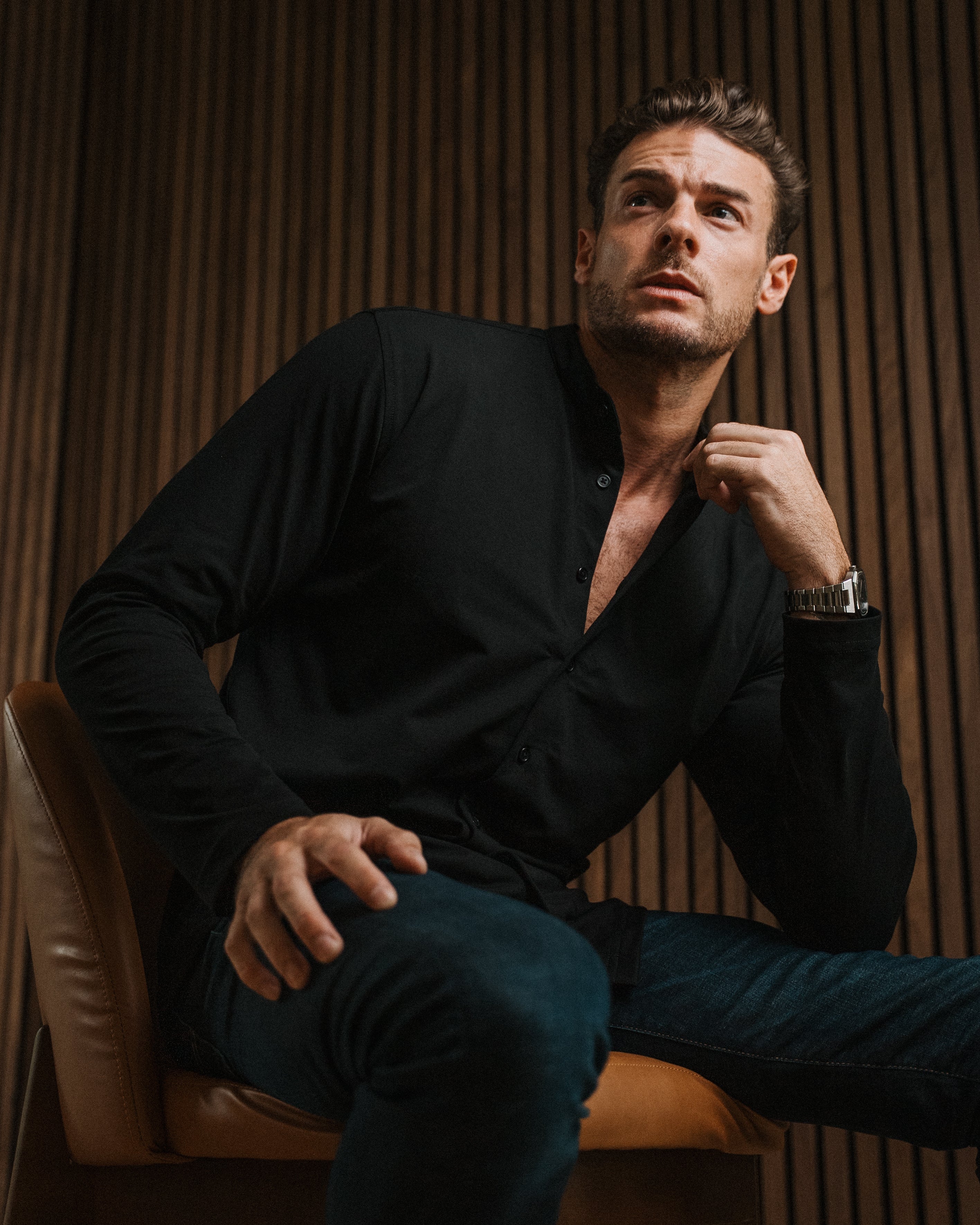 A man wearing a black long-sleeve button-up shirt and dark blue jeans sits on a brown leather chair against a dark wood slatted wall.