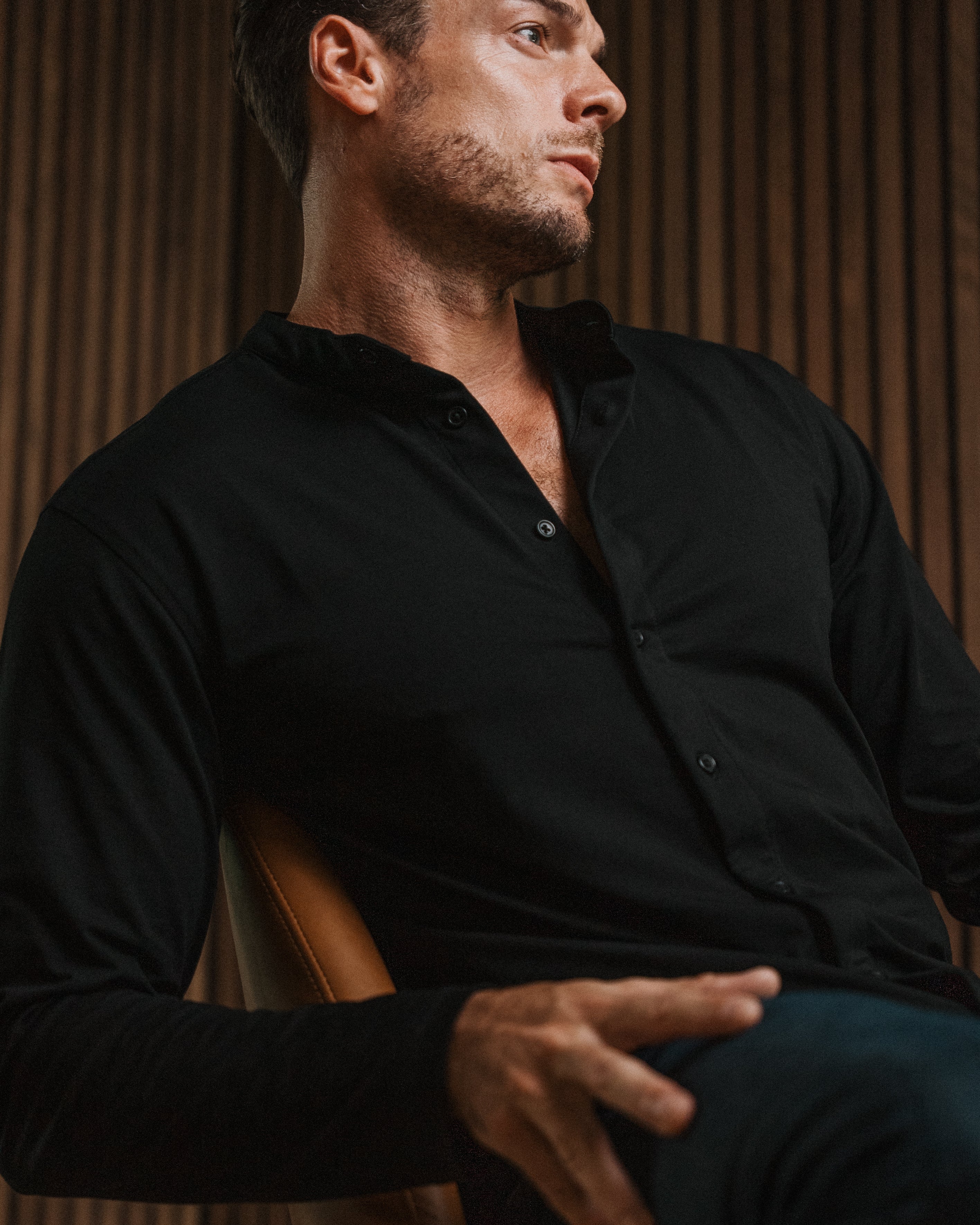 A man wearing a black long-sleeve button-up shirt and dark pants, seated in a leather chair against a warm wood slat wall.