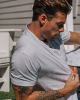 A man wearing a light gray short-sleeve polo shirt, shown from the chest up in an outdoor setting with a stone wall and house siding in the background.