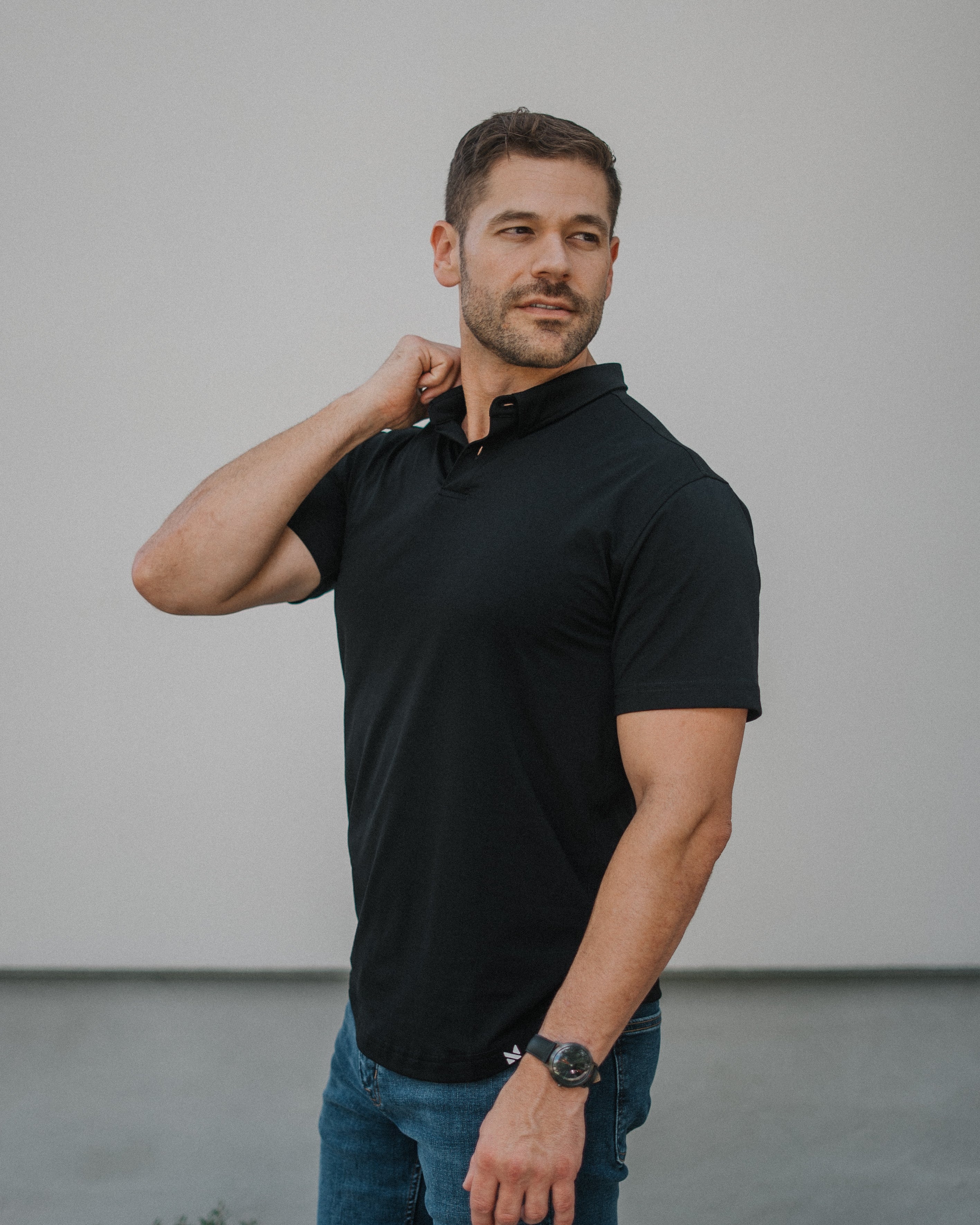 A man wearing a black short-sleeve polo shirt and blue jeans, standing against a light gray wall.