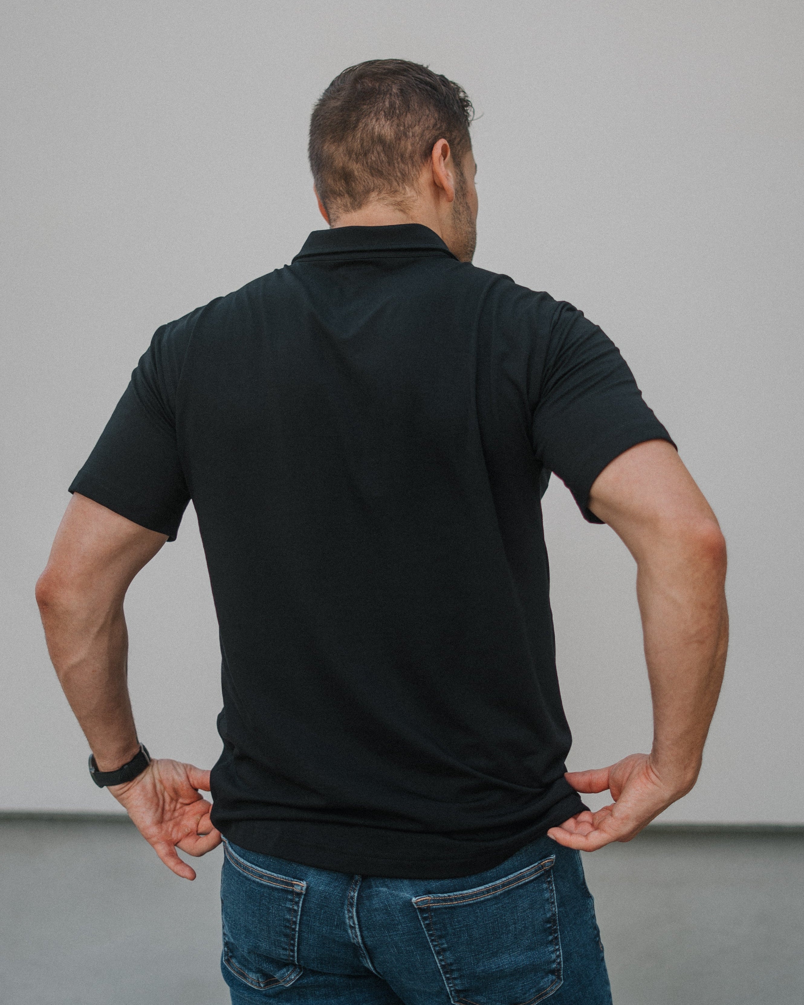 A man viewed from behind wearing a black short-sleeve polo shirt and blue jeans, standing against a plain light gray wall.