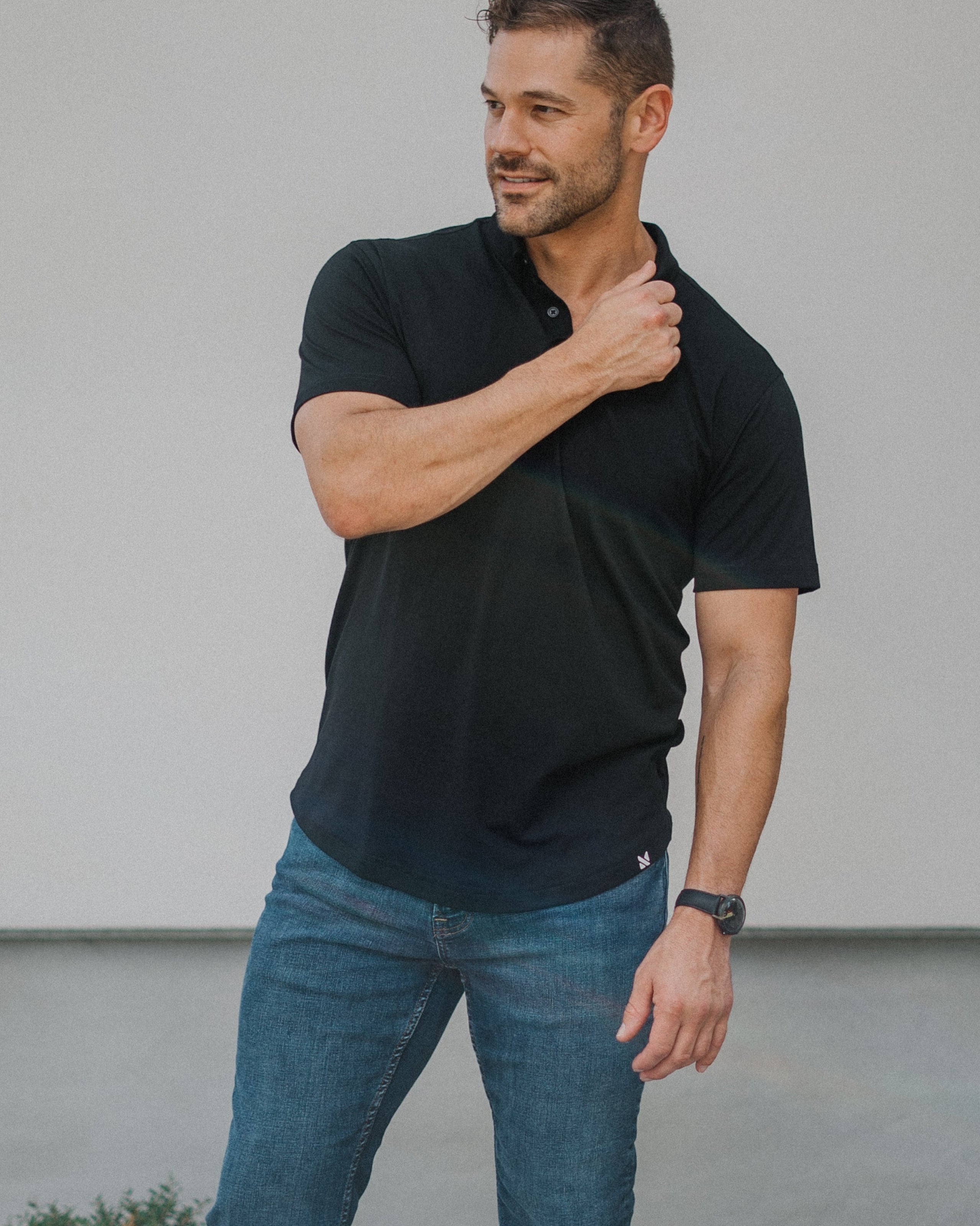 A man wearing a black short-sleeve polo shirt, blue jeans, and white sneakers stands outdoors against a light gray wall.
