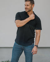 A man wearing a black short-sleeve polo shirt, blue jeans, and white sneakers stands outdoors against a light gray wall.