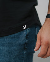 Close-up of a person wearing a black t-shirt and blue jeans, showing a small white geometric logo on the bottom hem of the shirt.