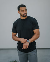 A man wearing a black short-sleeve crewneck t-shirt and gray pants, standing against a light gray wall.
