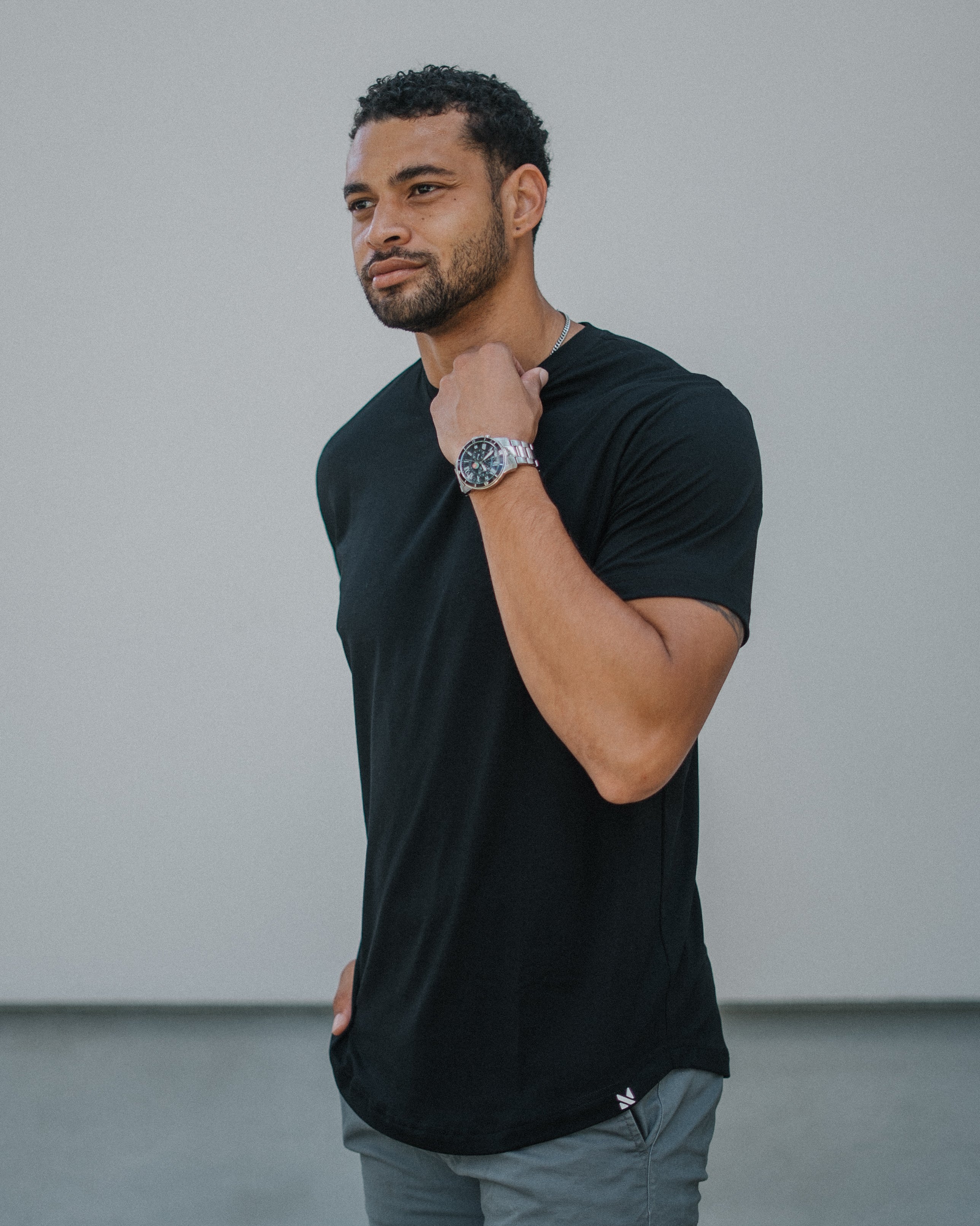 A man wearing a black short-sleeve athletic t-shirt and gray pants, standing against a neutral gray wall background.