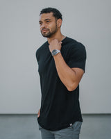 A man wearing a black short-sleeve athletic t-shirt and gray pants, standing against a neutral gray wall background.