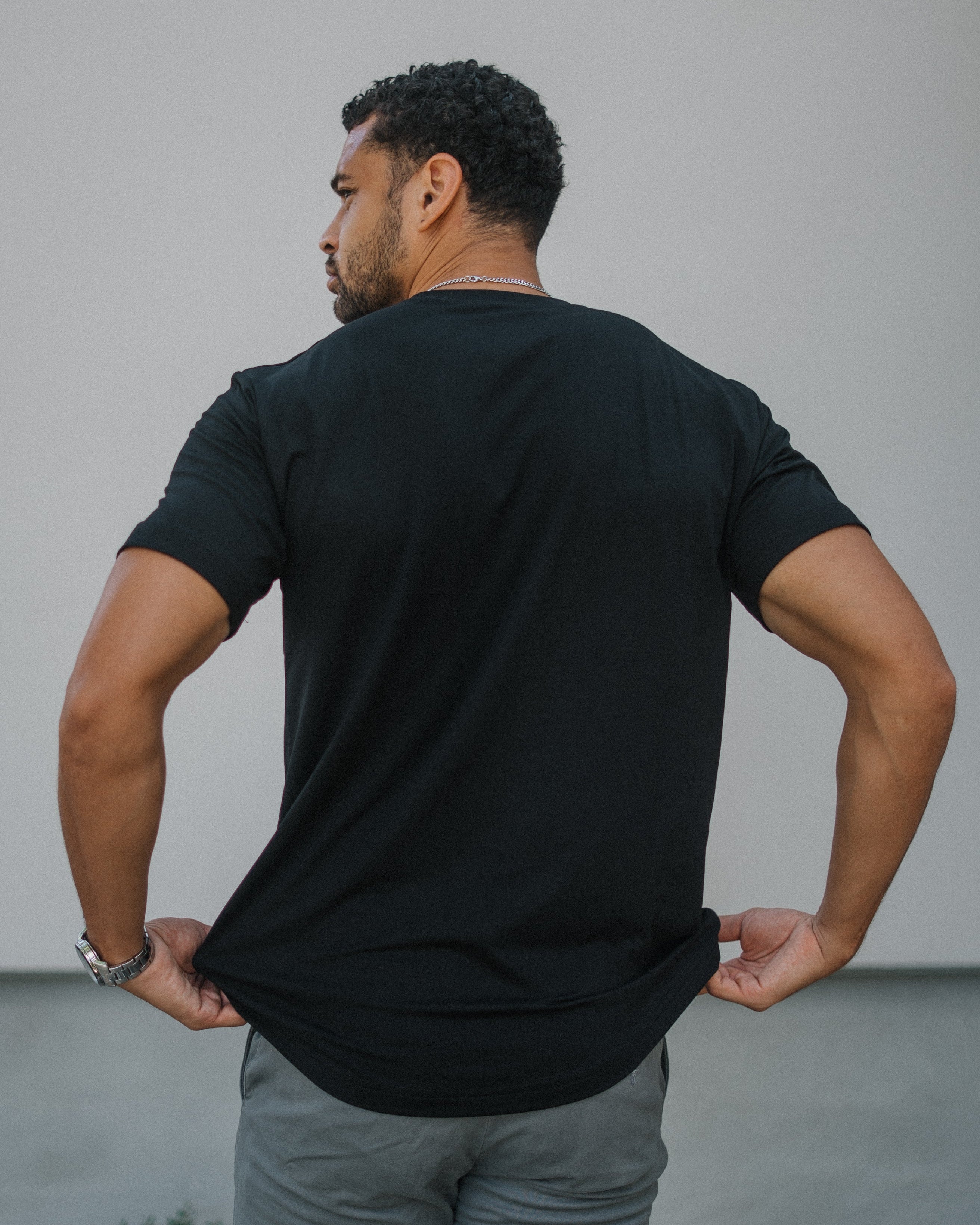 A man viewed from behind wearing a black short-sleeve t-shirt and gray pants, standing against a plain light gray wall.