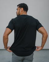 A man viewed from behind wearing a black short-sleeve t-shirt and gray pants, standing against a plain light gray wall.