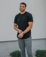 A man wearing a black crewneck t-shirt, gray slim-fit pants, and white sneakers, standing outdoors on a stone path in front of a modern light gray wall.