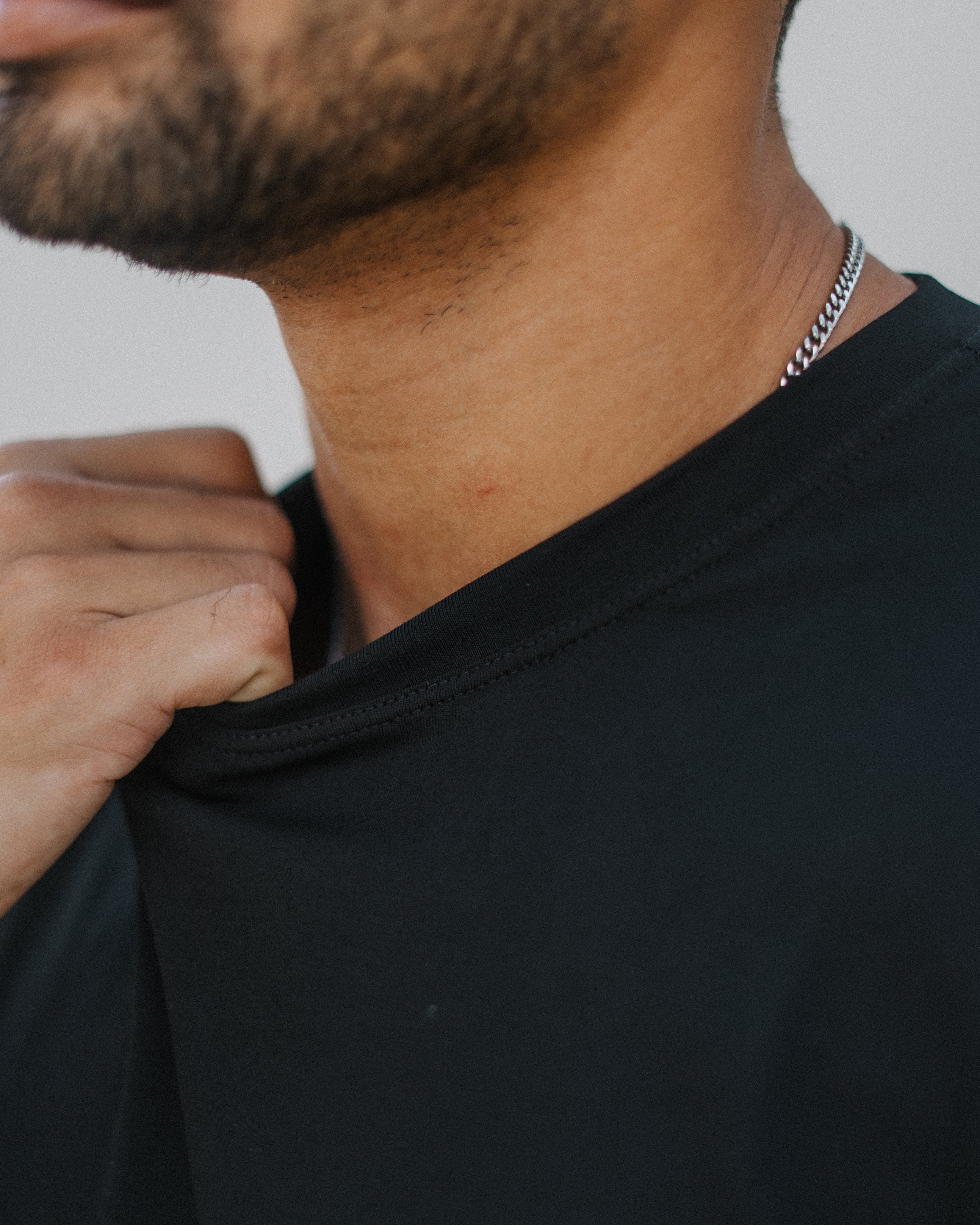 Close-up of a man wearing a black crew-neck t-shirt and a silver chain necklace, adjusting the collar of the shirt.