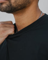 Close-up of a man wearing a black crew-neck t-shirt and a silver chain necklace, adjusting the collar of the shirt.