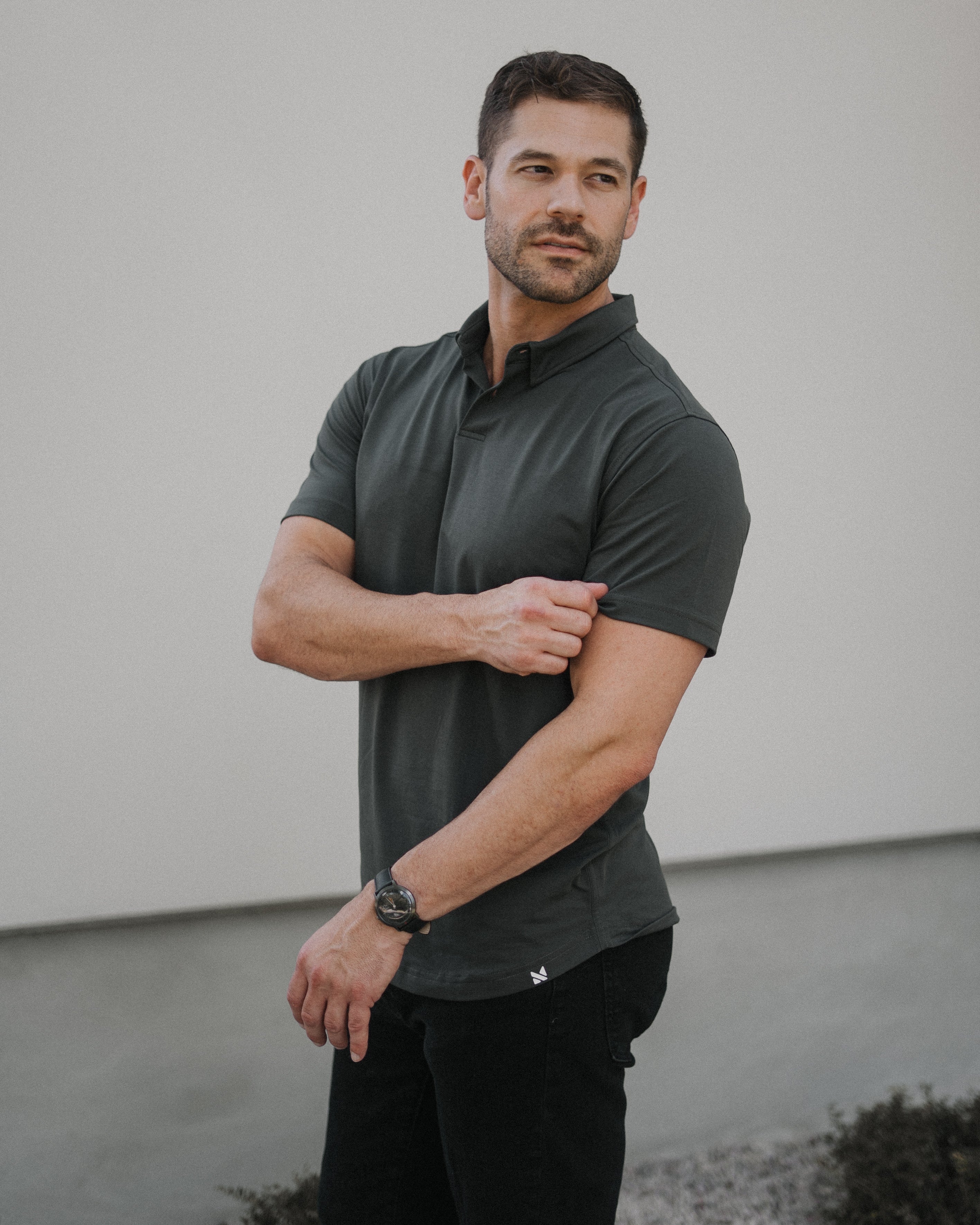 A man wearing a dark charcoal gray short-sleeve polo shirt and black pants, standing outdoors against a plain light gray wall.