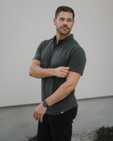 A man wearing a dark charcoal gray short-sleeve polo shirt and black pants, standing outdoors against a plain light gray wall.