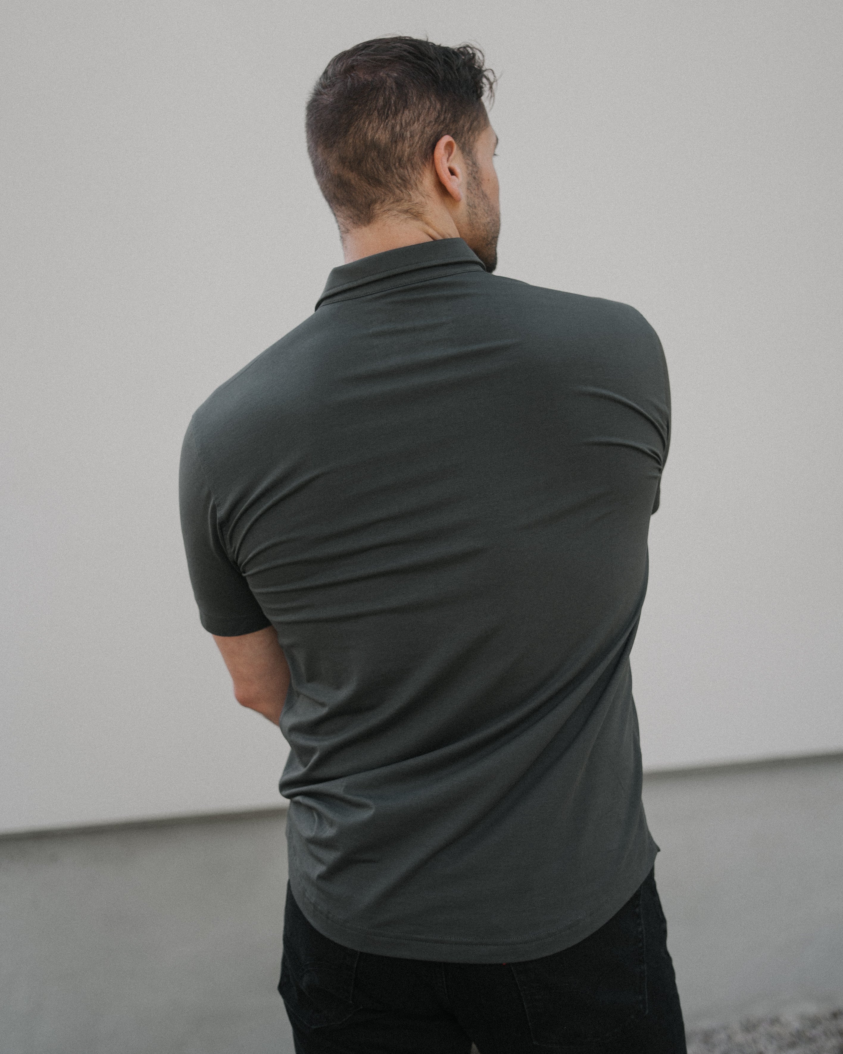 A man viewed from behind wearing a dark charcoal gray short-sleeve polo shirt and black pants, standing against a minimalist light gray wall.