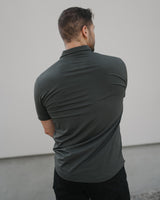 A man viewed from behind wearing a dark charcoal gray short-sleeve polo shirt and black pants, standing against a minimalist light gray wall.