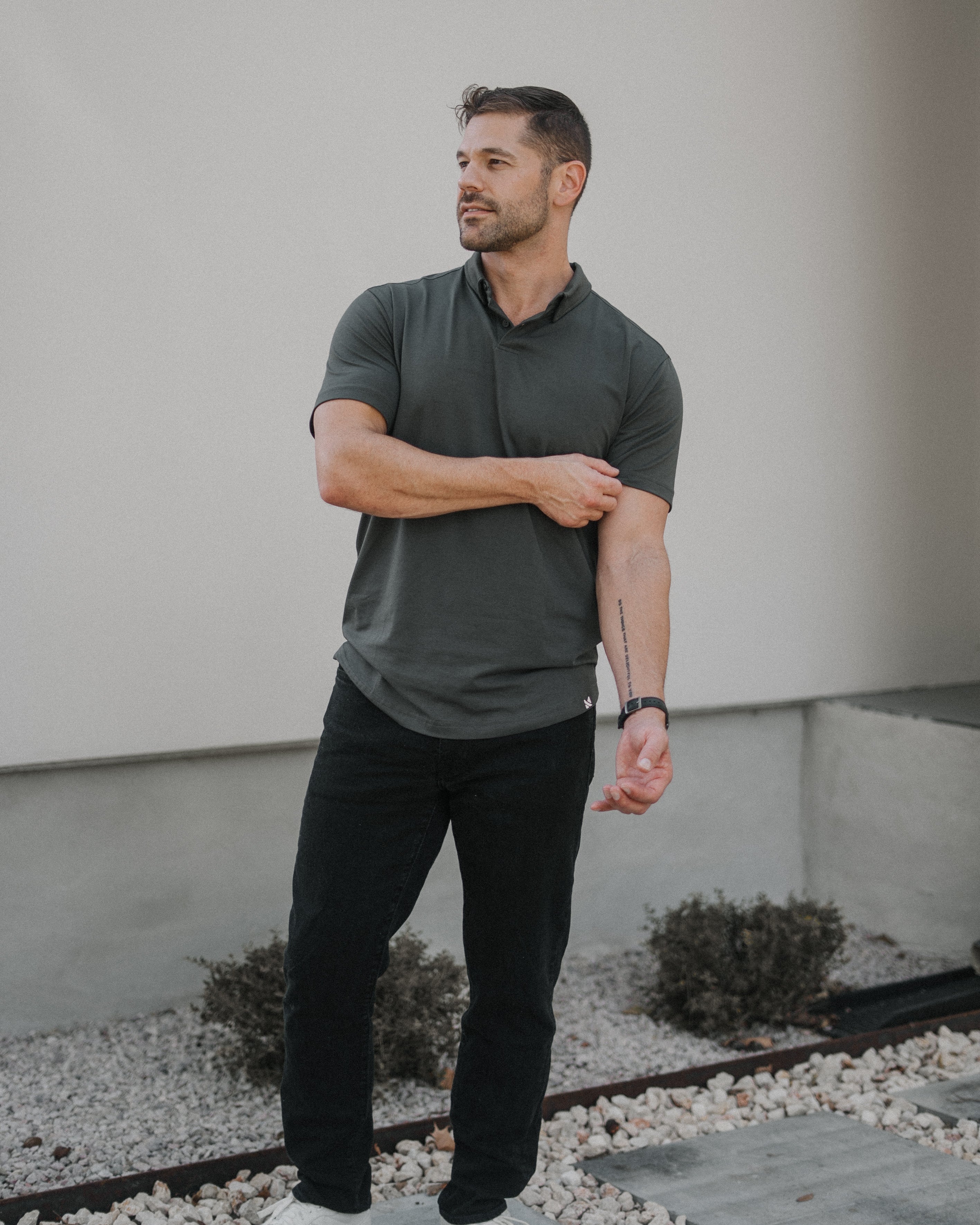 A man wearing a dark gray short-sleeve polo shirt, black pants, and white sneakers standing outdoors in front of a light-colored wall.