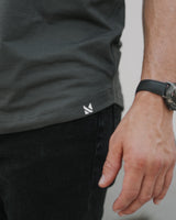 Close-up of a person wearing a dark charcoal gray athletic t-shirt with a small white geometric logo near the hem, paired with black pants and a black watch.