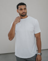 A man wearing a plain white short-sleeve crewneck t-shirt and gray pants, standing against a light-colored wall.