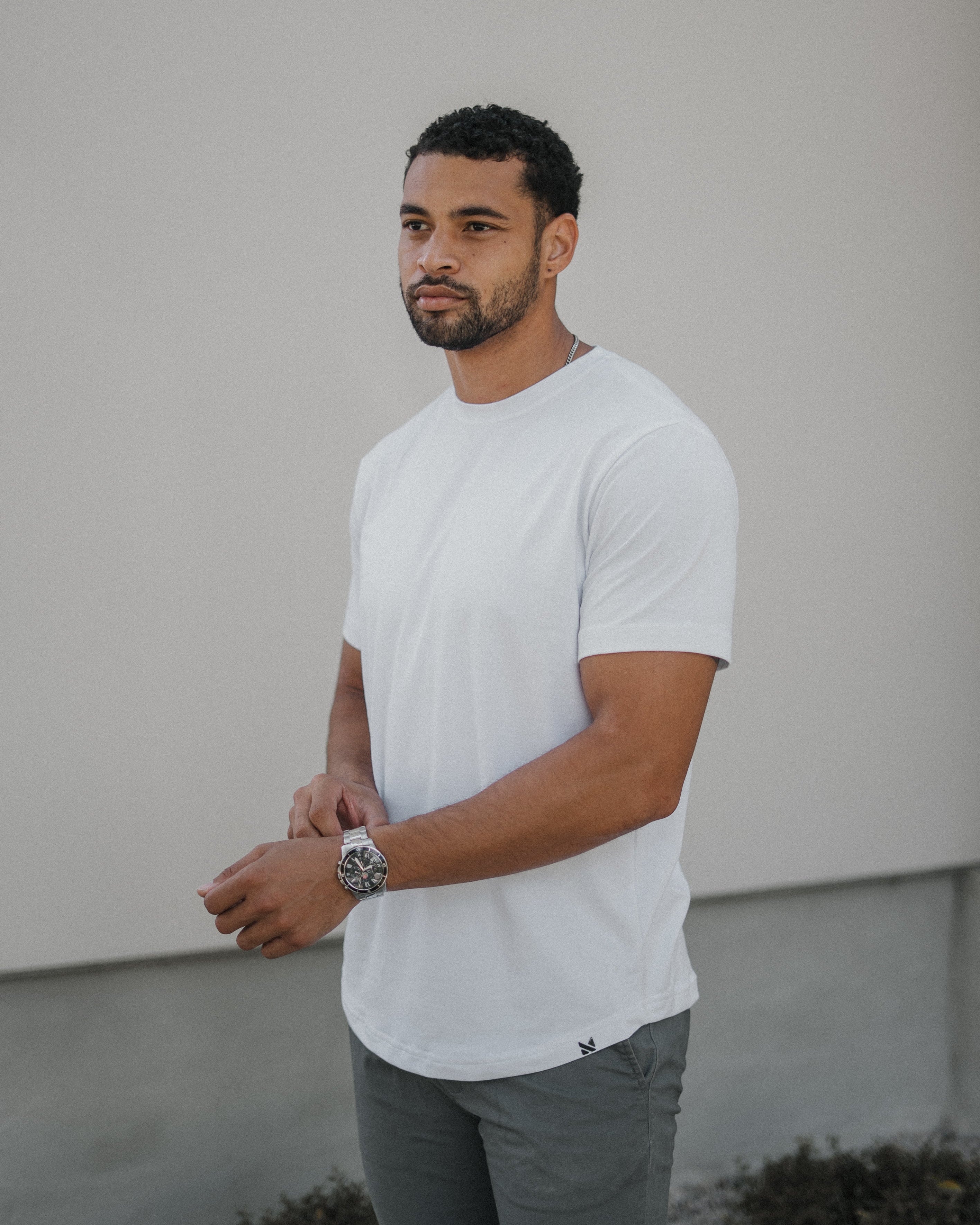 A man wearing a plain white short-sleeve crewneck t-shirt and gray pants, standing against a light gray stucco wall.