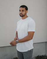 A man wearing a plain white short-sleeve crewneck t-shirt and gray pants, standing against a light gray stucco wall.