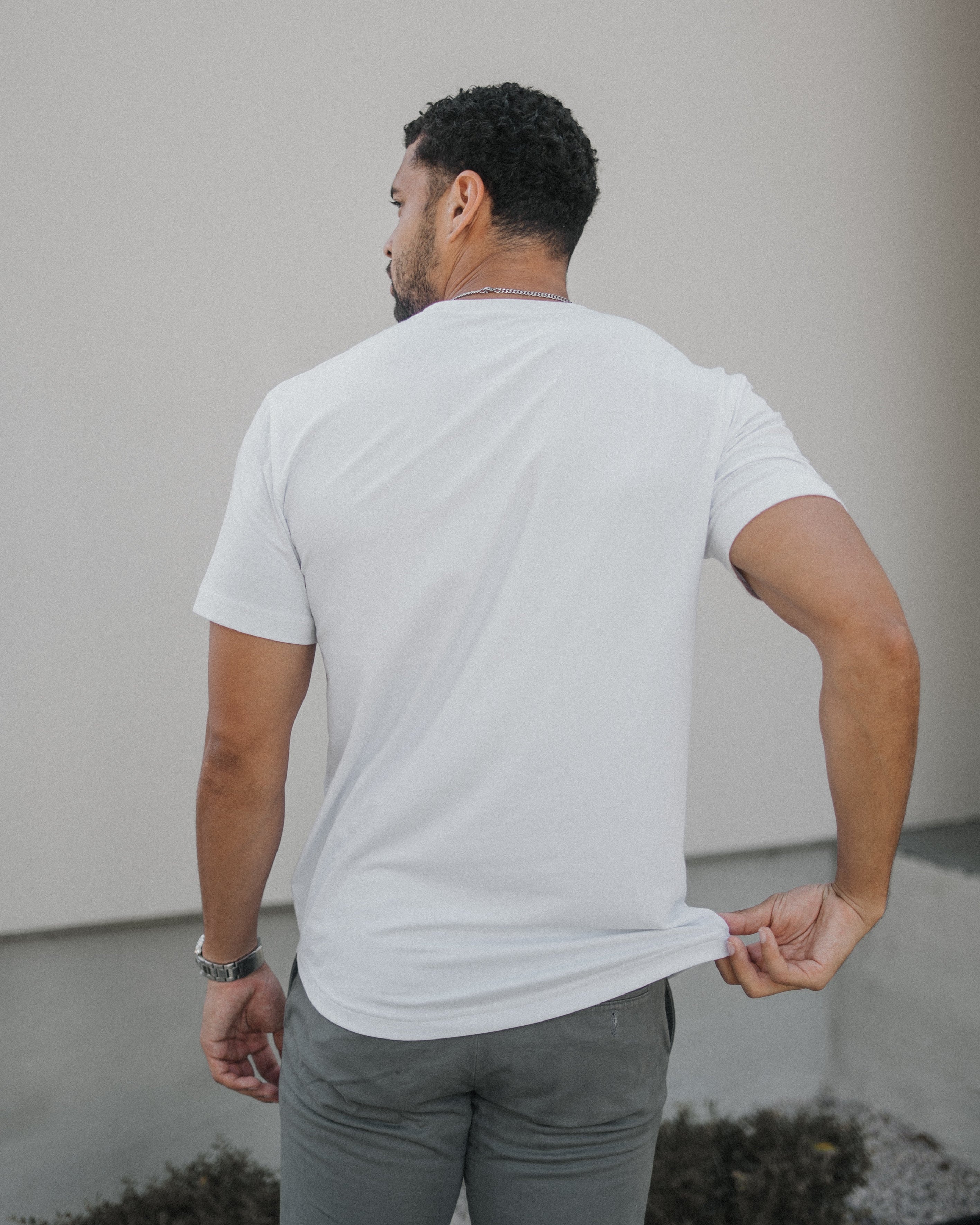 A man viewed from behind wearing a plain white short-sleeve t-shirt and gray pants, standing outdoors against a light-colored wall.