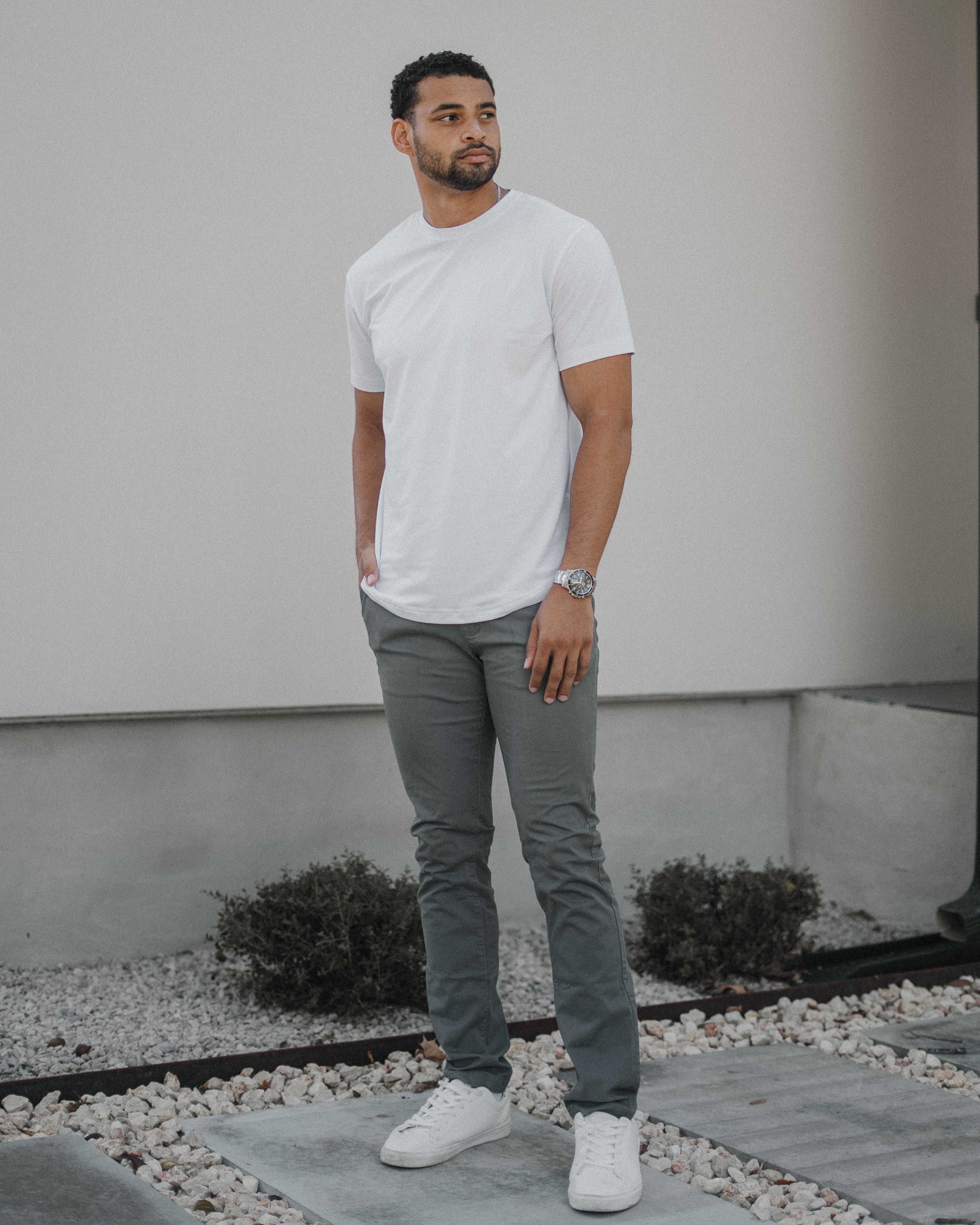 A man wearing a white crew-neck t-shirt, gray slim-fit jeans, and white sneakers stands outdoors in front of a modern house wall.