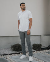 A man wearing a white crew-neck t-shirt, gray slim-fit jeans, and white sneakers stands outdoors in front of a modern house wall.