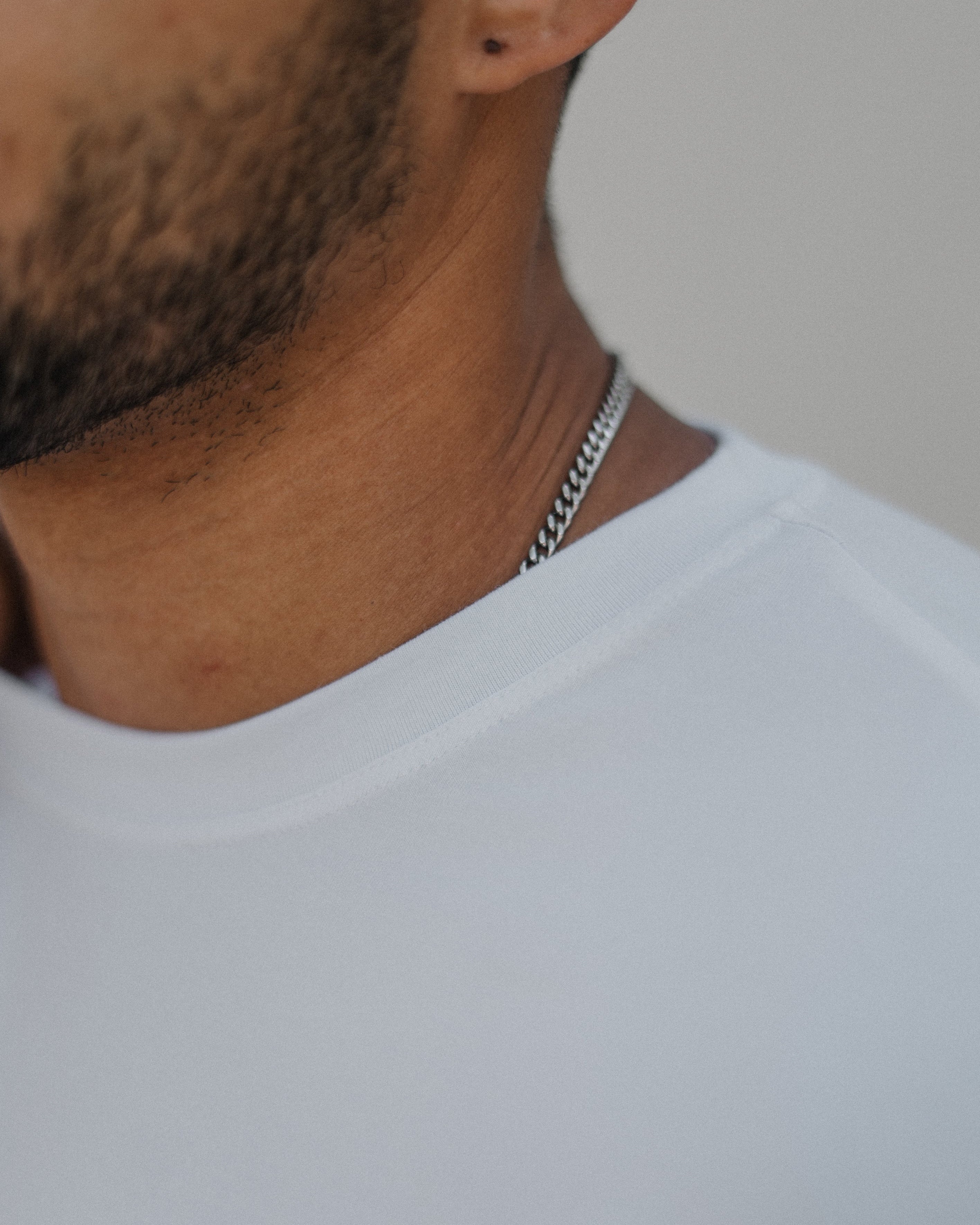 A close-up shot of a man's neck and upper chest wearing a silver chain necklace and a white t-shirt against a neutral light gray background.
