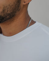 A close-up shot of a man's neck and upper chest wearing a silver chain necklace and a white t-shirt against a neutral light gray background.