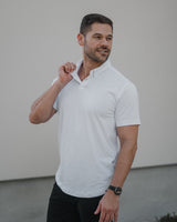 A man wearing a white short-sleeve polo shirt and black pants, standing against a minimalist light gray wall.
