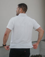 A man viewed from behind wearing a white short-sleeve polo shirt and black jeans, standing against a neutral light gray wall.
