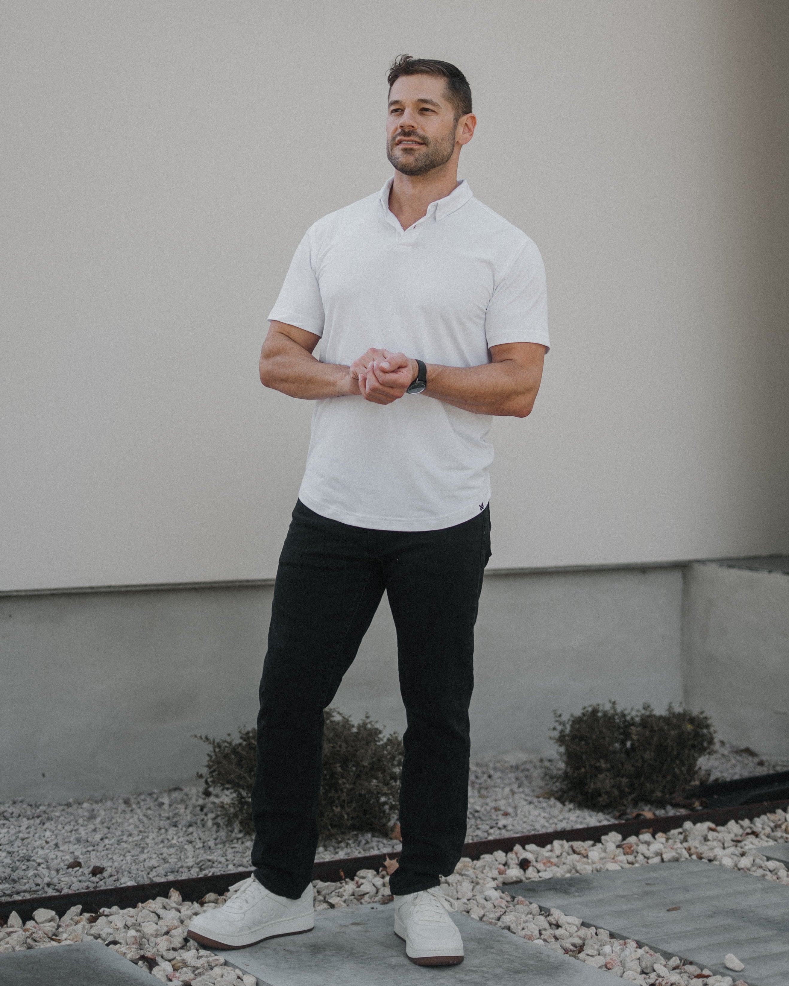 A man wearing a white short-sleeve polo shirt, black pants, and white sneakers stands outdoors in front of a light-colored stucco wall.
