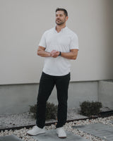 A man wearing a white short-sleeve polo shirt, black pants, and white sneakers stands outdoors in front of a light-colored stucco wall.