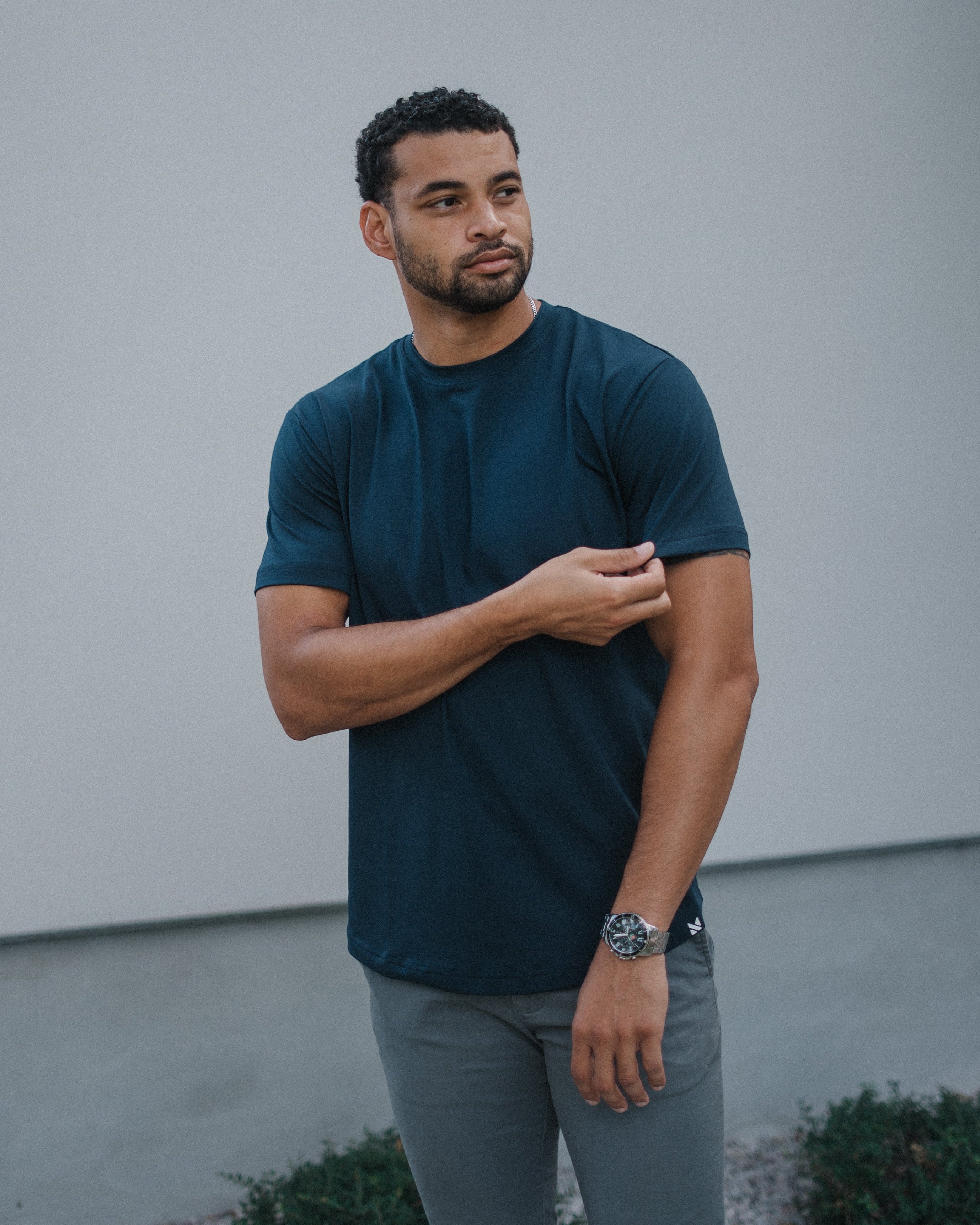 A man wearing a dark navy blue short-sleeve crewneck t-shirt and gray pants, standing outdoors against a light gray wall.