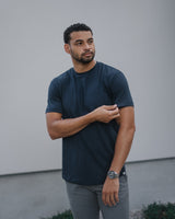 A man wearing a dark navy blue crewneck t-shirt and gray pants, standing outdoors against a light gray wall.
