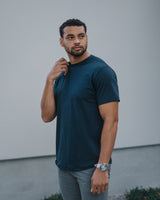 A man wearing a dark navy blue crewneck t-shirt and gray pants, standing outdoors against a light gray wall.