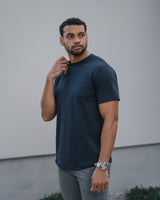 A man wearing a dark navy blue crewneck t-shirt and gray pants, standing outdoors against a light gray wall.