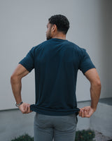 A man viewed from behind wearing a dark navy blue short-sleeve t-shirt and gray pants, pulling the hem of the shirt slightly outward with both hands against a modern light gray building wall.