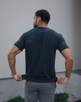 A man viewed from behind wearing a dark navy blue t-shirt and gray pants, pulling the hem of the shirt slightly with both hands in front of a modern light gray building wall.