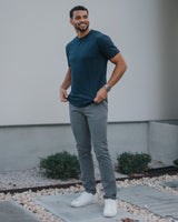 A man wearing a dark teal crewneck t-shirt, gray slim-fit pants, and white sneakers, standing outdoors in front of a modern white wall.