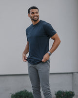 A man wearing a dark teal crewneck t-shirt, gray slim-fit pants, and white sneakers, standing outdoors in front of a modern white wall.