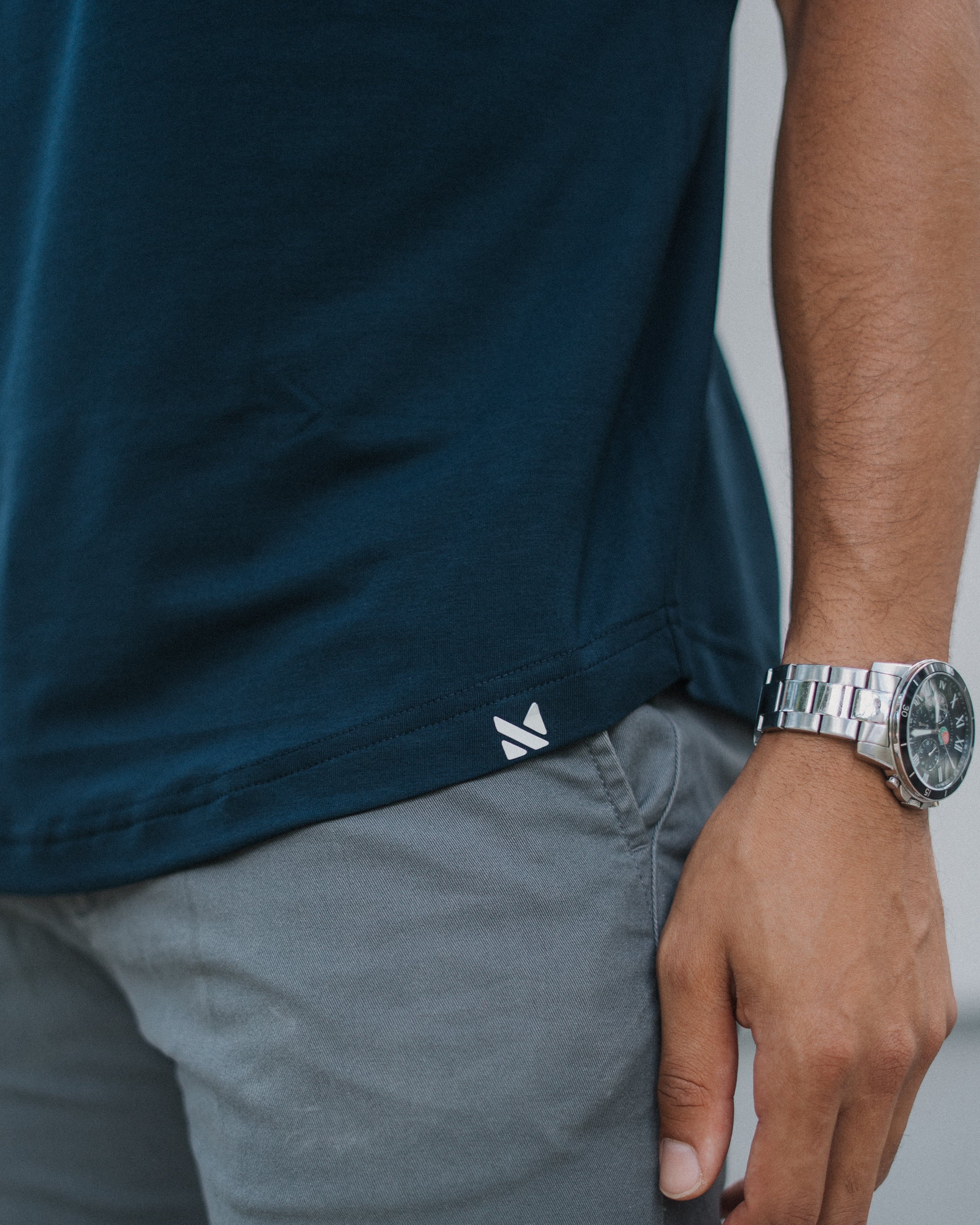 Close-up of a person wearing a dark teal short-sleeve t-shirt and gray pants, with a silver watch on the wrist. A small white geometric logo is visible near the hem of the shirt.