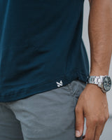 Close-up of a person wearing a dark teal short-sleeve t-shirt and gray pants, with a silver watch on the wrist. A small white geometric logo is visible near the hem of the shirt.