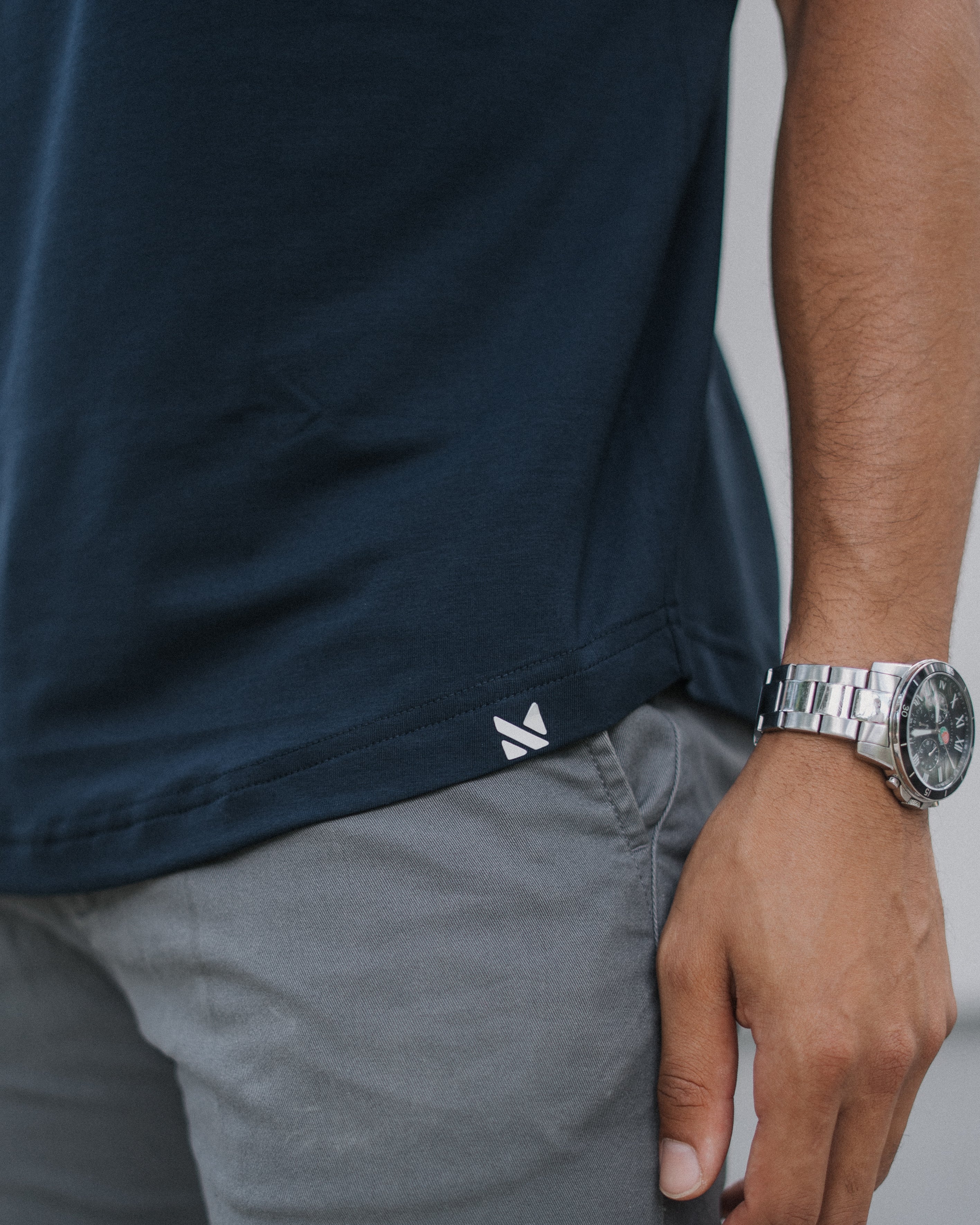 Close-up of a person wearing a dark navy short-sleeve t-shirt and gray pants, with a silver watch on the wrist. A small white logo is visible near the hem of the shirt.