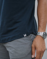 Close-up of a person wearing a dark navy short-sleeve t-shirt and gray pants, with a silver watch on the wrist. A small white logo is visible near the hem of the shirt.