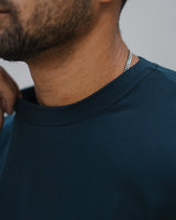Close-up of a man wearing a dark navy blue crewneck t-shirt and a silver chain necklace, with his neck and lower face visible against a neutral gray background.