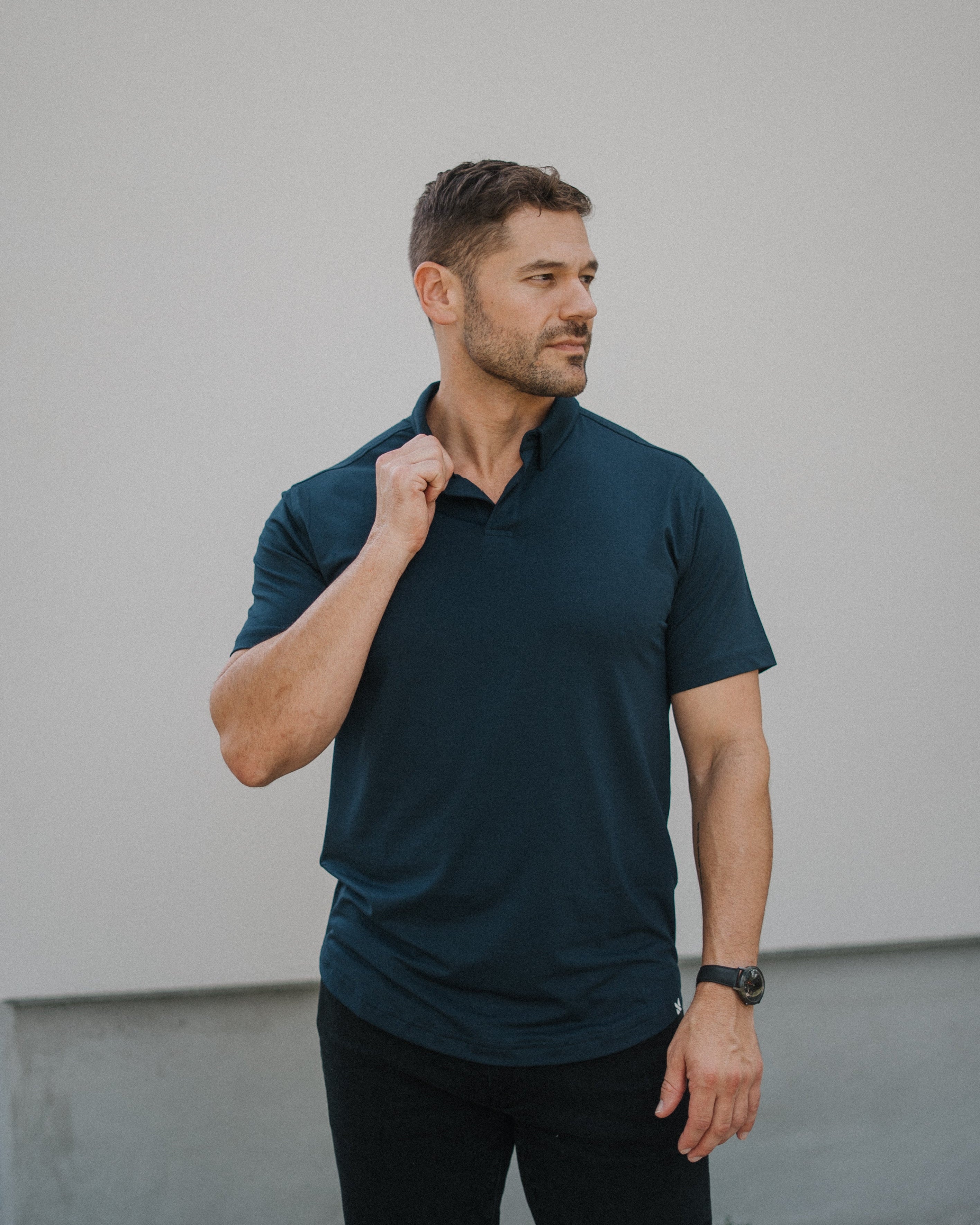A man wearing a dark navy blue short-sleeve polo shirt and black pants, standing against a light gray wall.