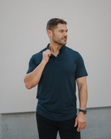 A man wearing a dark navy blue short-sleeve polo shirt and black pants, standing against a light gray wall.