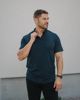 A man wearing a dark navy blue short-sleeve polo shirt and black pants, standing against a light gray wall.